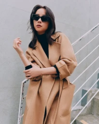 Model wearing dark sunglasses and a long camel wool coat posing on outdoor stairs with a white railing for Max Mara.