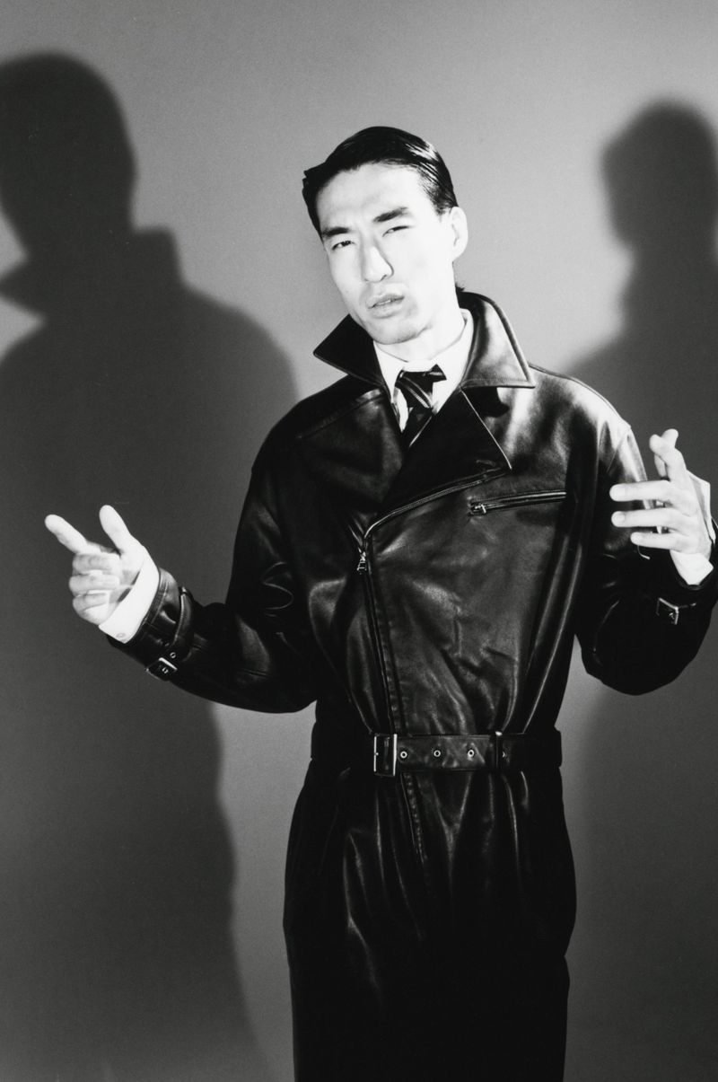 Monochrome portrait of a man wearing a black leather coat against a grey studio backdrop with prominent shadows.