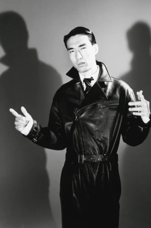 Monochrome portrait of a man wearing a black leather coat against a grey studio backdrop with prominent shadows.