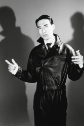 Monochrome portrait of a man wearing a black leather coat against a grey studio backdrop with prominent shadows.