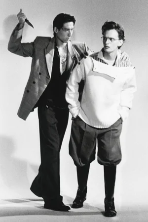 Black and white editorial image of two male models posing, one holding a knife behind the other in a studio setting.