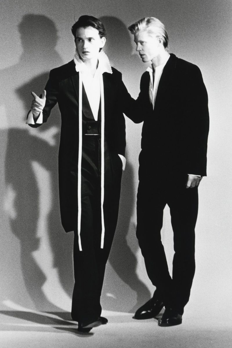 Black and white editorial image of two male models in dark suits standing against a light seamless studio backdrop.