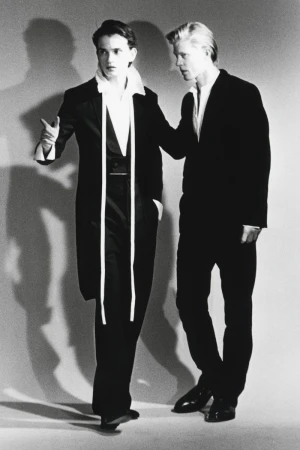 Black and white editorial image of two male models in dark suits standing against a light seamless studio backdrop.