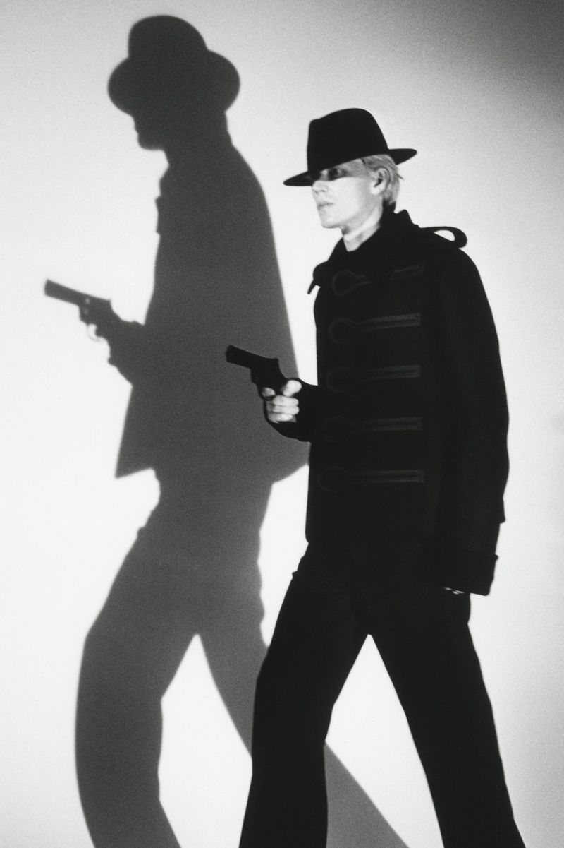 Model in dark fedora hat and black coat holding a handgun with a sharp dramatic shadow on the wall for HERO Magazine.
