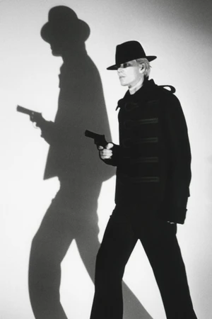 Model in dark fedora hat and black coat holding a handgun with a sharp dramatic shadow on the wall for HERO Magazine.