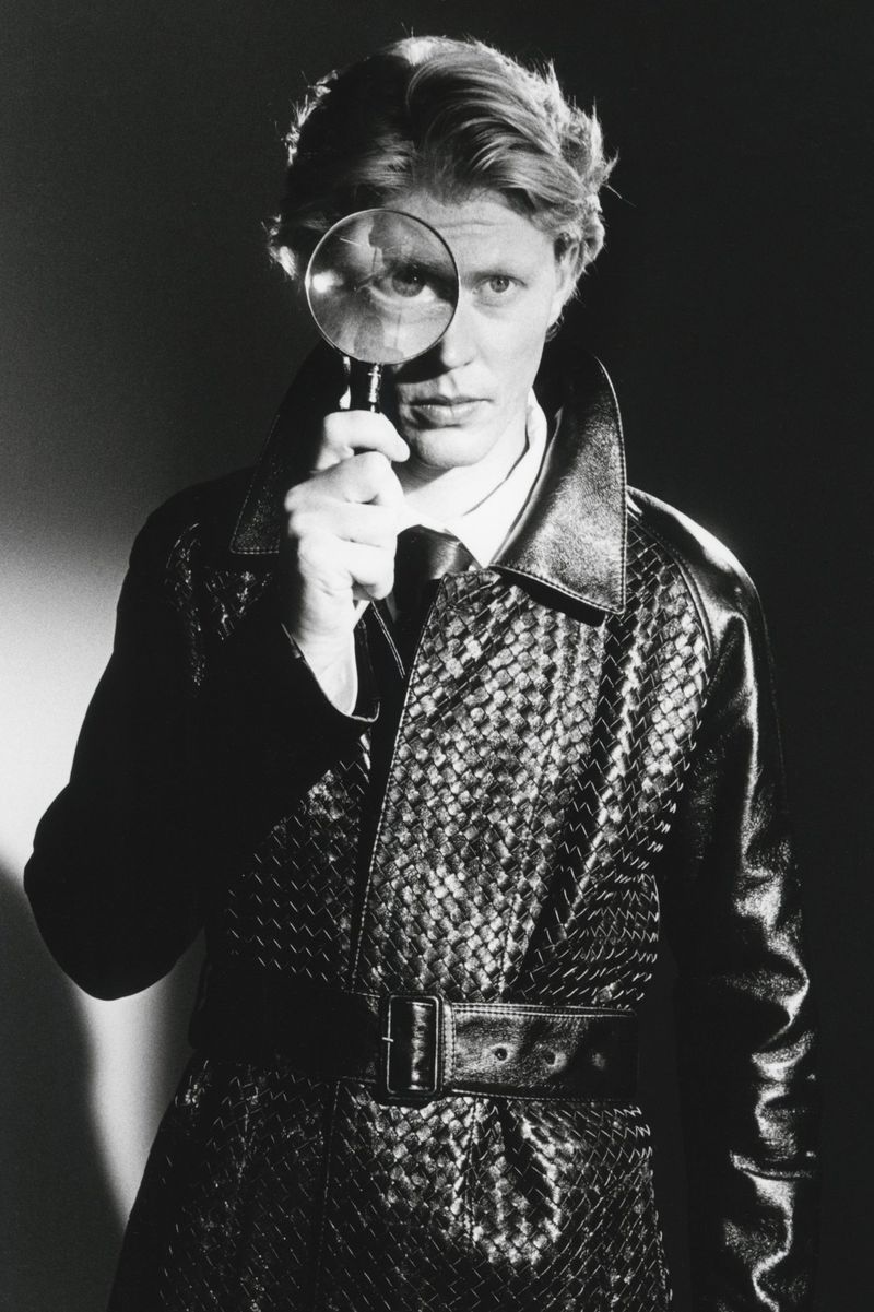 Man wearing a textured black leather trench coat holding up a magnifying glass in front of his eye for HERO Magazine.