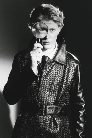 Man wearing a textured black leather trench coat holding up a magnifying glass in front of his eye for HERO Magazine.