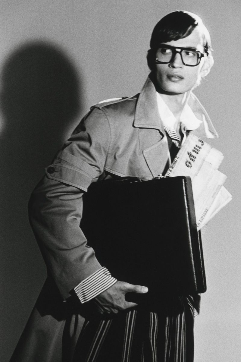 Man wearing glasses and trench coat carrying a briefcase and newspapers in a black and white editorial photograph.