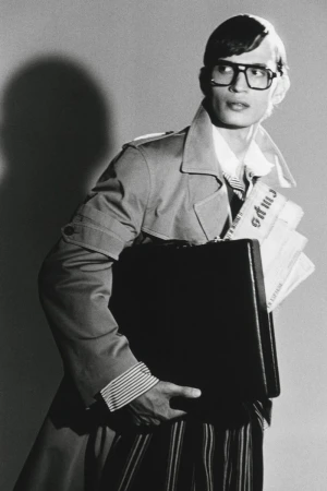 Man wearing glasses and trench coat carrying a briefcase and newspapers in a black and white editorial photograph.