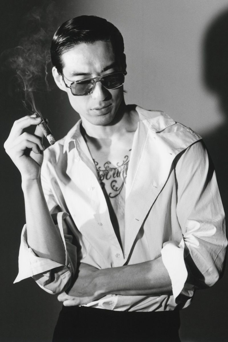 Monochrome portrait of a man wearing sunglasses holding a lit cigar in front of a plain background.