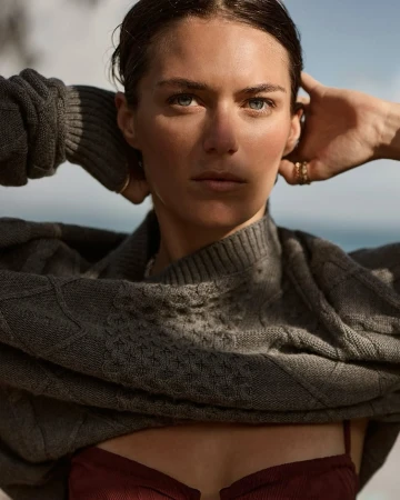 Woman with blue eyes and dark hair poses in a gray knit sweater and reddish-brown top with a sea and bright sky background.