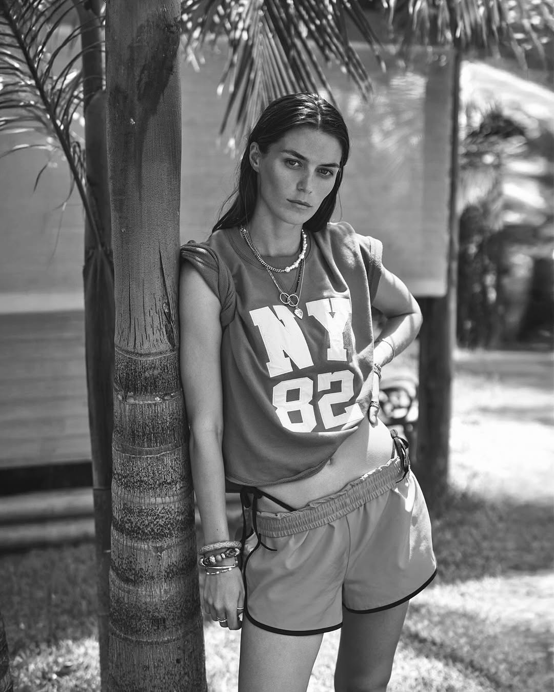Black and white portrait of a woman in shorts and a tee shirt leaning against a palm tree outdoors with filtered sunlight.