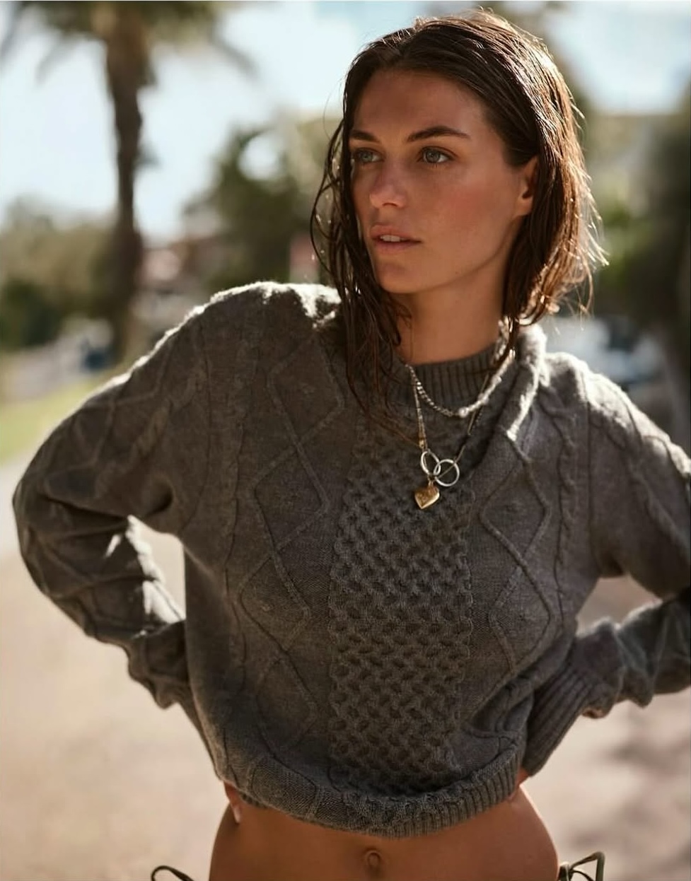 Close-up of an adult woman with wet hair wearing a gray sweater and layered necklaces, posing outdoors in natural sunlight.