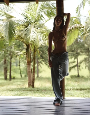 Adult female model posing against a wooden post on a porch with palm trees and green foliage in the background on a sunny day.