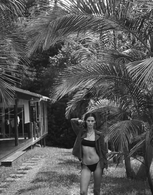 Monochrome shot of a woman in a bikini and jacket standing in a tropical setting with palm trees and a rustic house in the background.