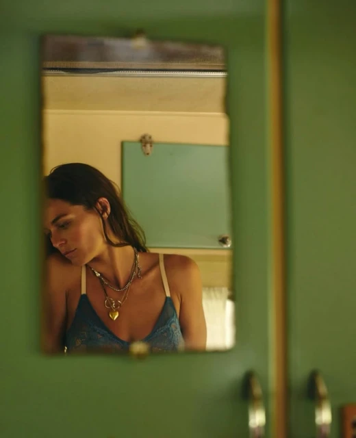 Reflection of a woman with dark hair and a blue top in a green-framed mirror, adorned with a gold heart necklace.