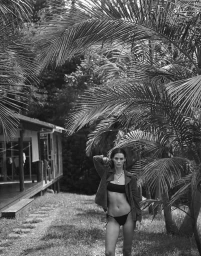 Model in a bikini top and jacket posing outdoors surrounded by lush palm fronds and a building.