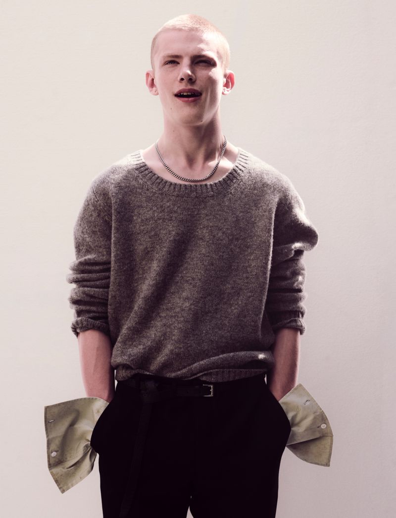 Young male model with a shaved head wearing a grey knit sweater and black trousers against a neutral background.