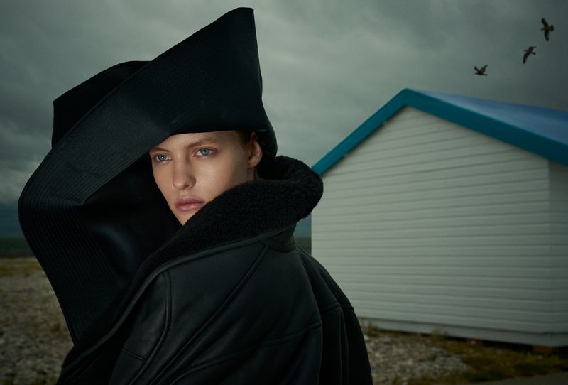 Model wearing a black sculptural hat and leather jacket outdoors under an overcast sky for a Numéro France editorial project.