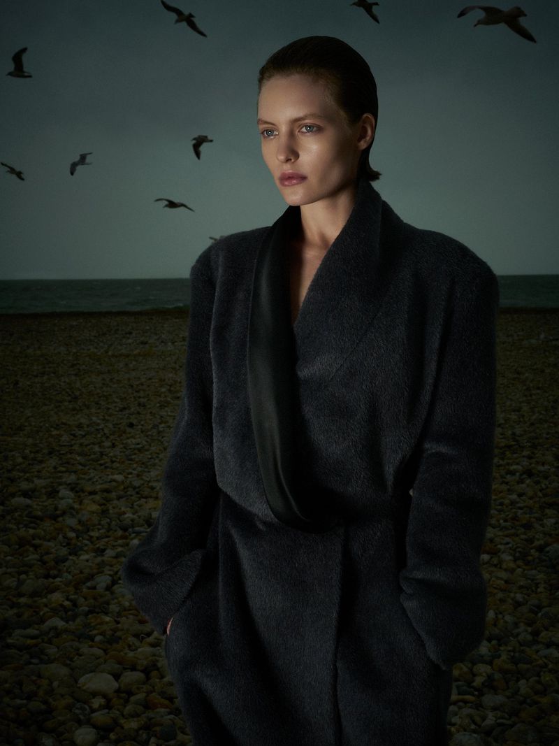Model in a dark charcoal coat standing on a shingle beach under an overcast sky with flying seabirds.