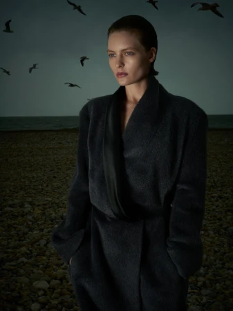 Model in a dark charcoal coat standing on a shingle beach under an overcast sky with flying seabirds.