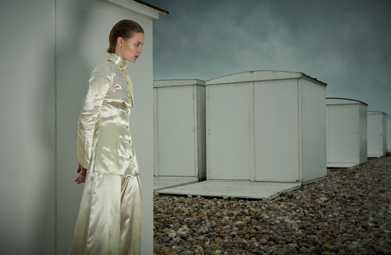 Model in shiny cream silk tunic and trousers standing near white beach cabins under stormy sky