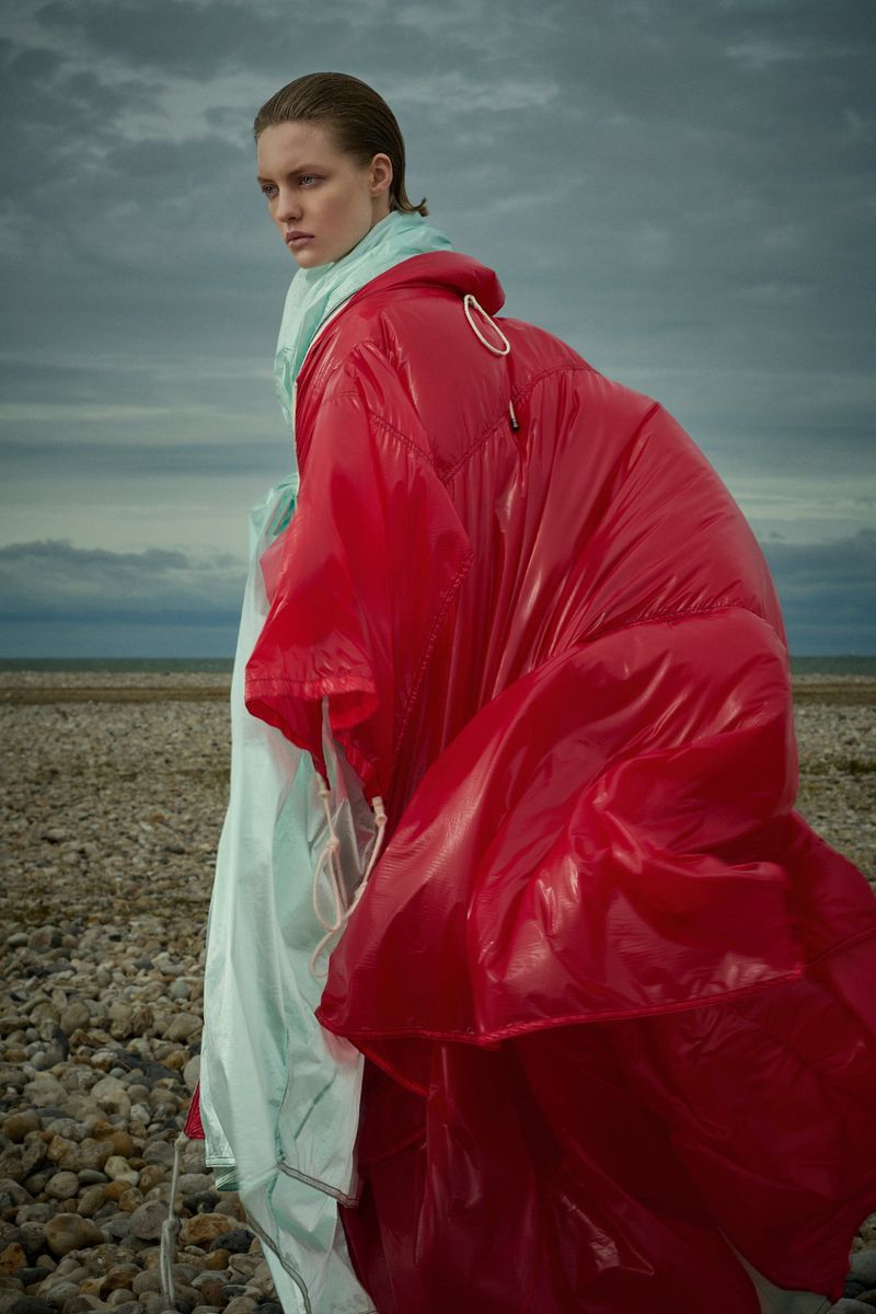 Model wearing a glossy red oversized puffer jacket and light mint scarf standing on a stony beach beneath a stormy grey sky