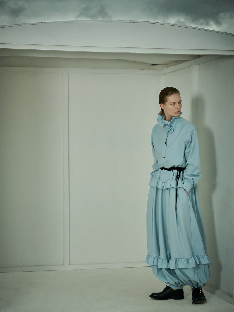 Model in blue ruffled dress and dark shoes standing against a plain white studio backdrop with stormy sky visible above.
