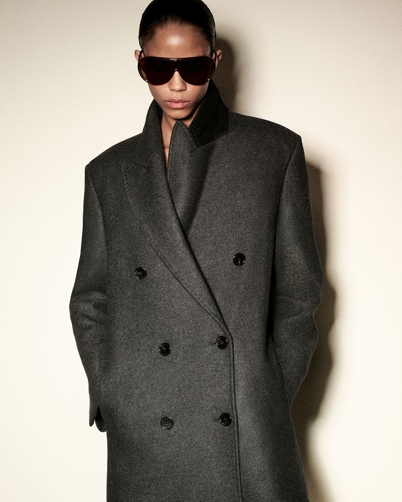Model in dark sunglasses wearing a charcoal double-breasted coat against a plain pale background