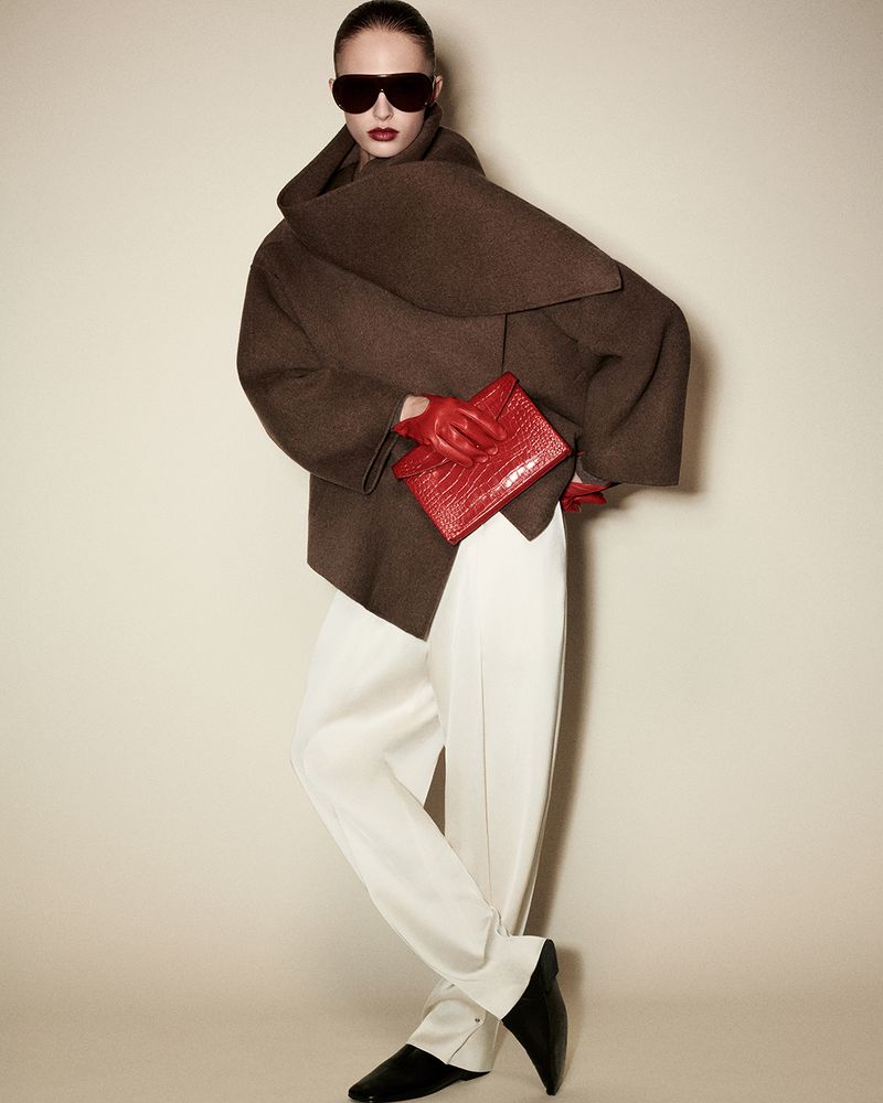 Model in oversized brown coat wearing dark sunglasses and holding a red clutch against a neutral background
