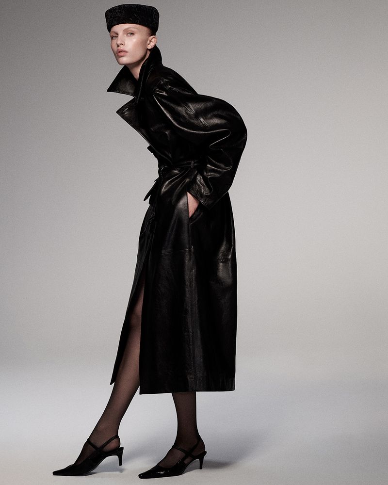 Model in black leather coat and pillbox hat posing against a neutral grey studio backdrop