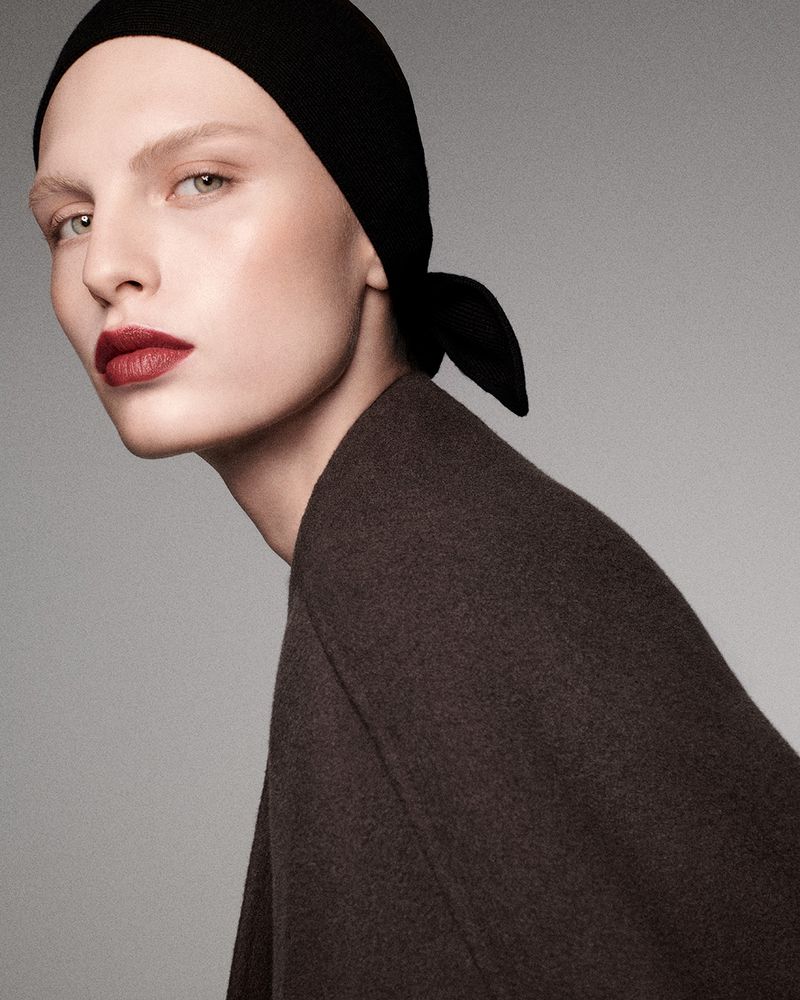 Close-up portrait of a model wearing a dark head covering and brown coat against a neutral grey studio background.
