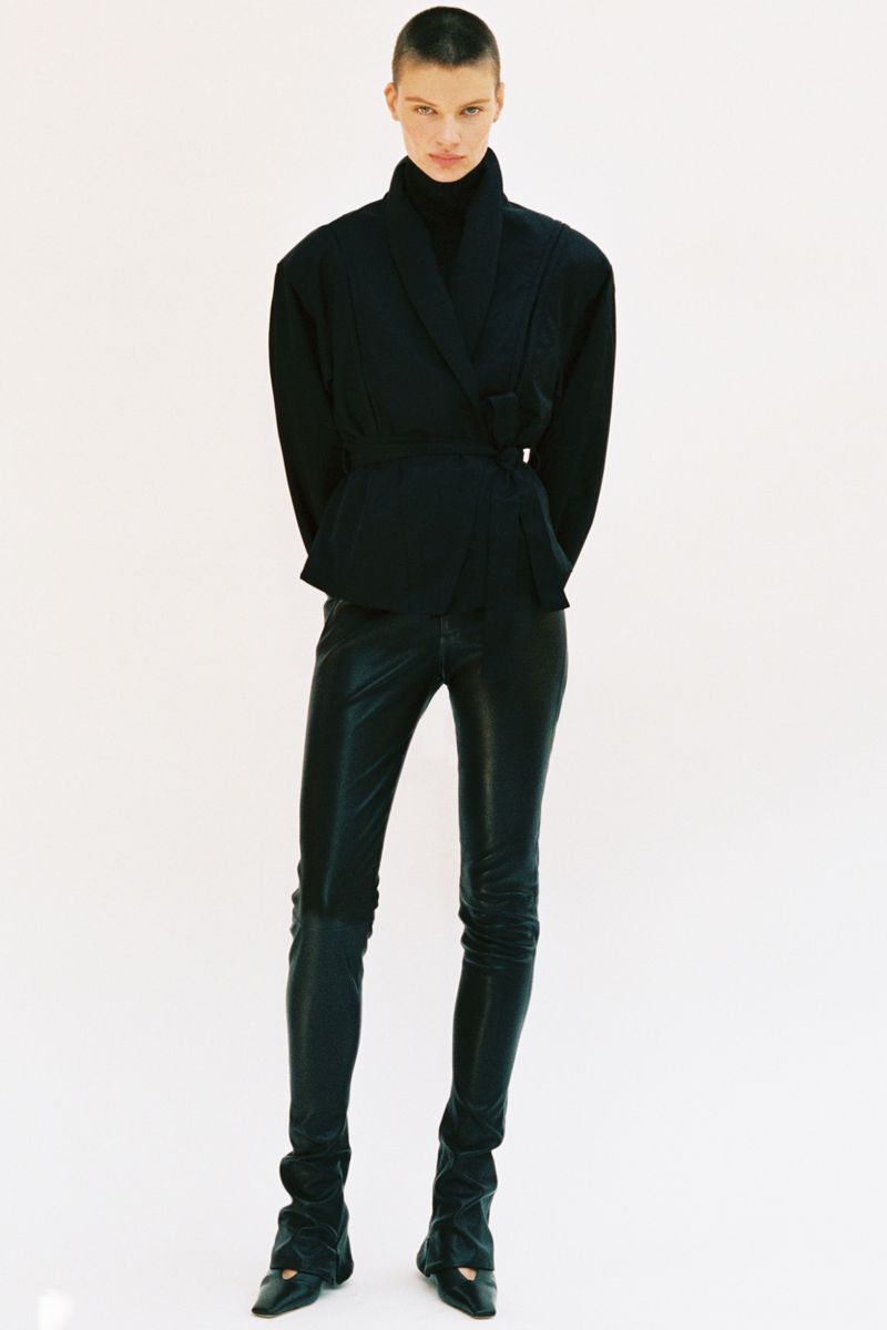 Model standing against a plain white background wearing a dark wrap jacket and dark trousers