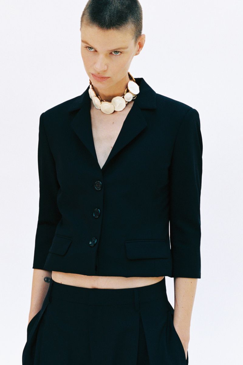 Model with short hair wearing a black buttoned blazer and a large round beaded necklace against a white background