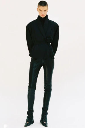 Model standing against a plain white background wearing a dark wrap jacket and dark trousers