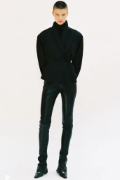 Model standing against a plain white background wearing a dark wrap jacket and dark trousers
