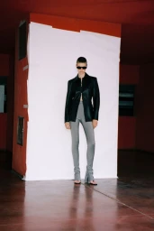 Model standing in dark sunglasses, black jacket, and fitted grey trousers against a white backdrop for Armarium.