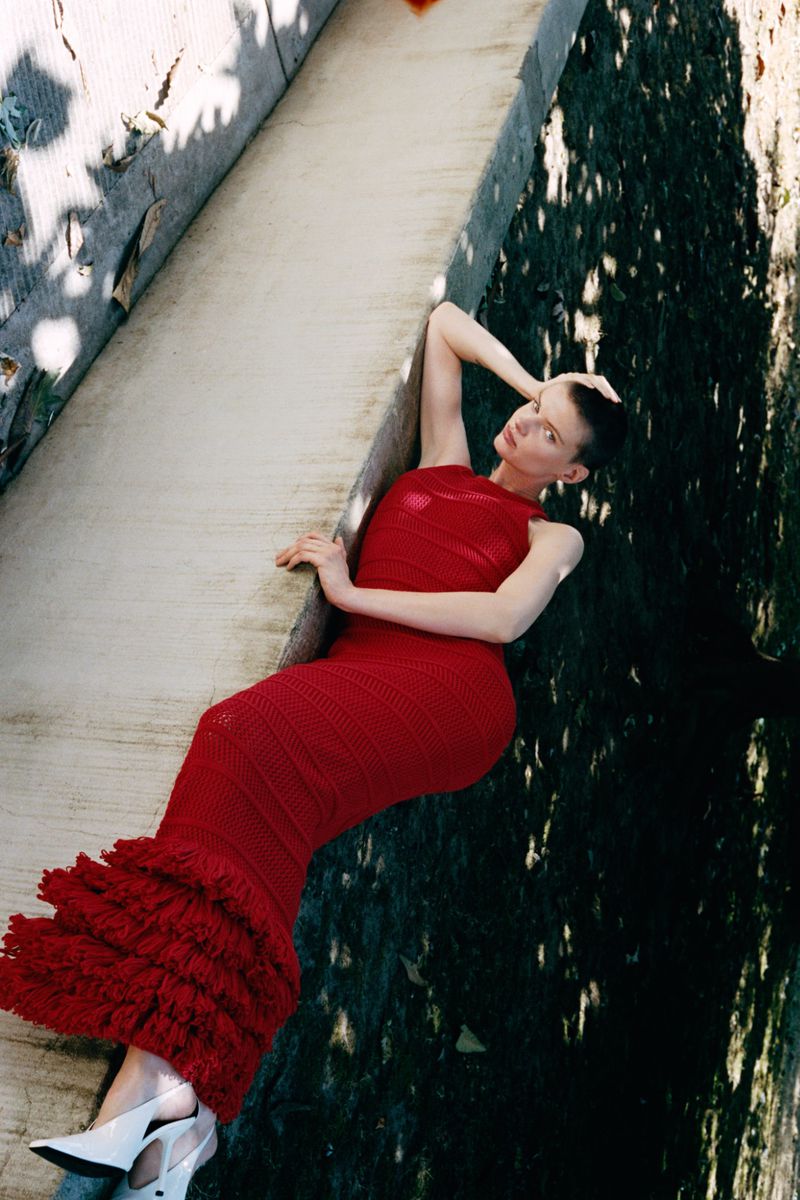 Model with a buzzcut wearing a long red knitted dress lying against a concrete wall outdoors