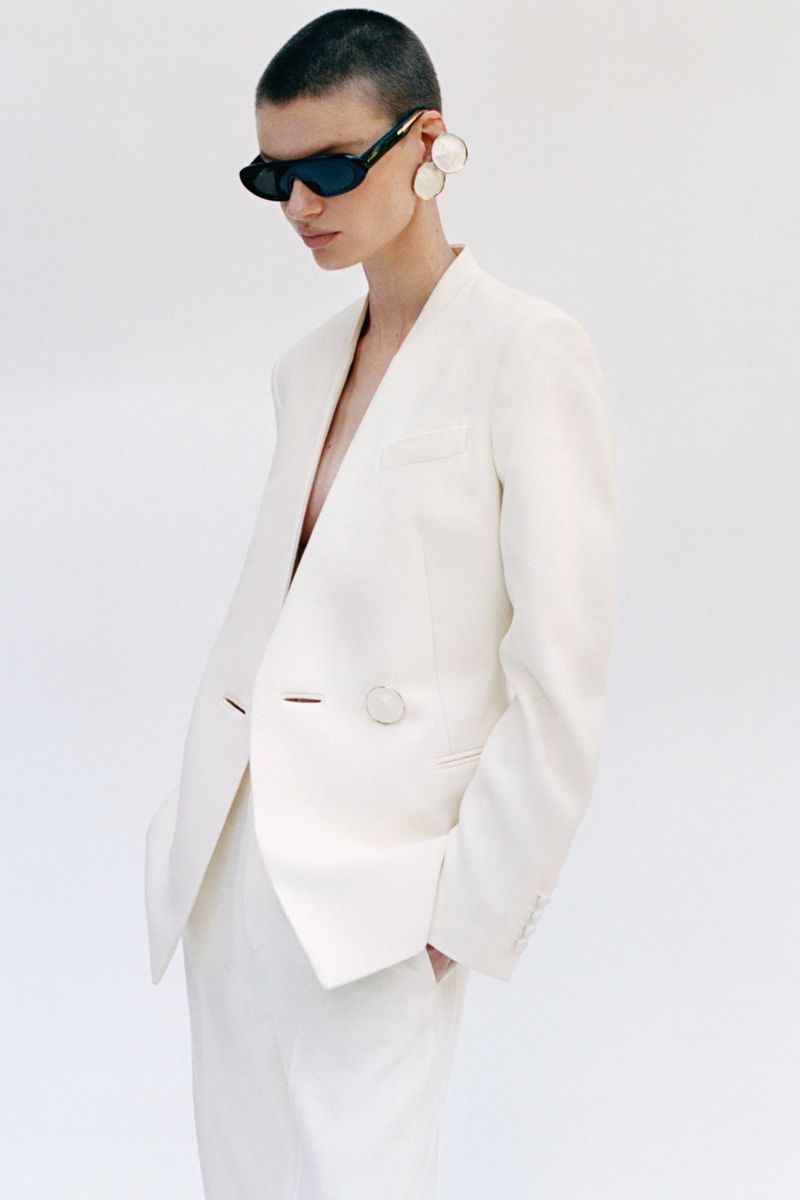 Model with a shaved head wearing a white blazer, dark sunglasses, and large statement earrings against a plain white background.