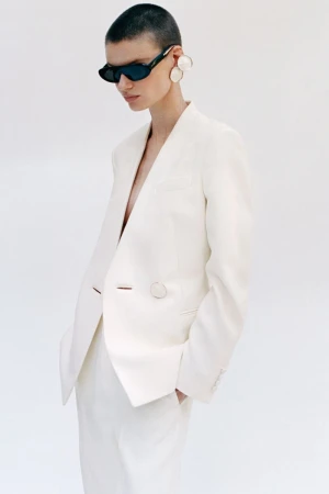 Model with a shaved head wearing a white blazer, dark sunglasses, and large statement earrings against a plain white background.