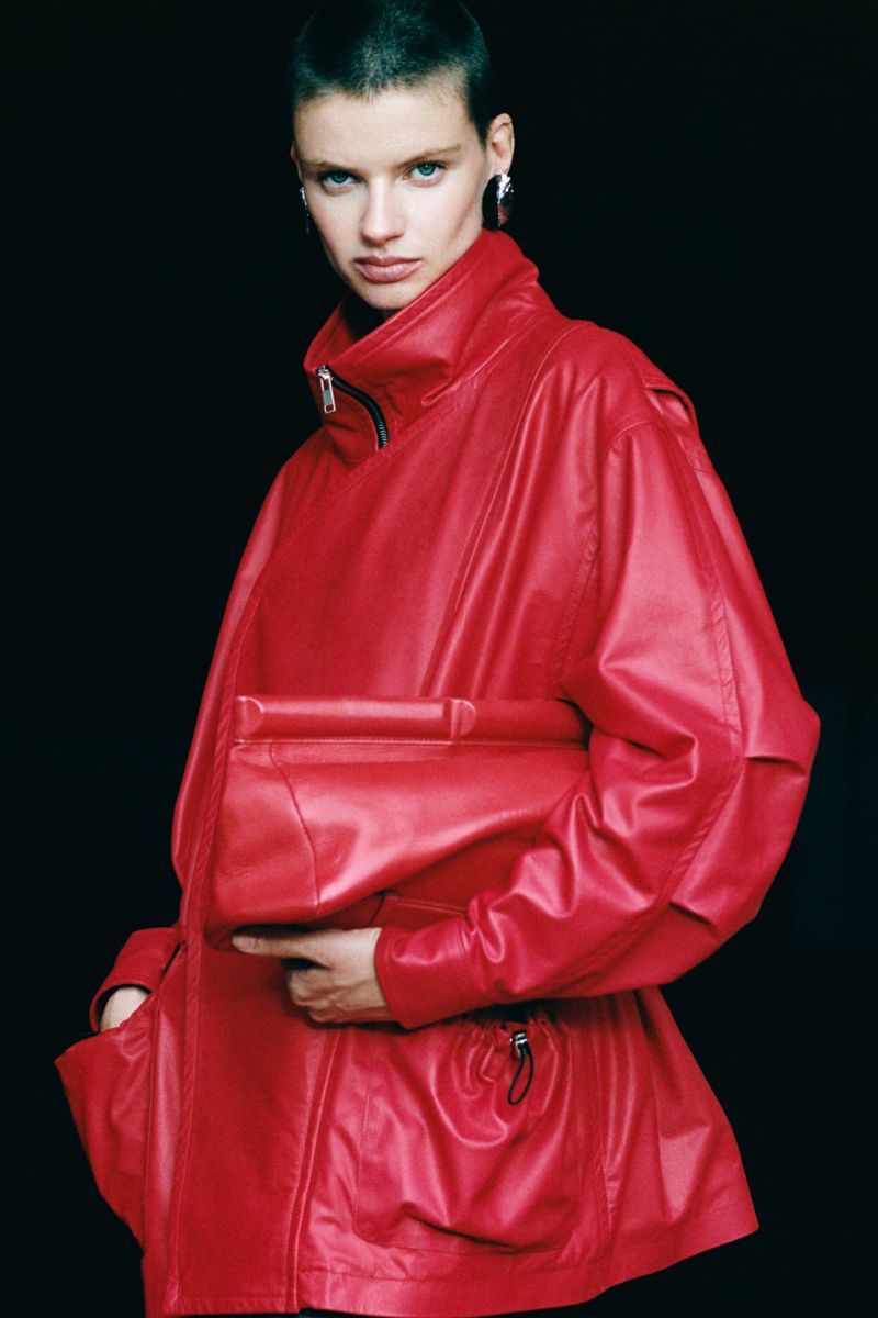 Model with short hair wearing a bright red leather jacket holding a matching red clutch against a black background