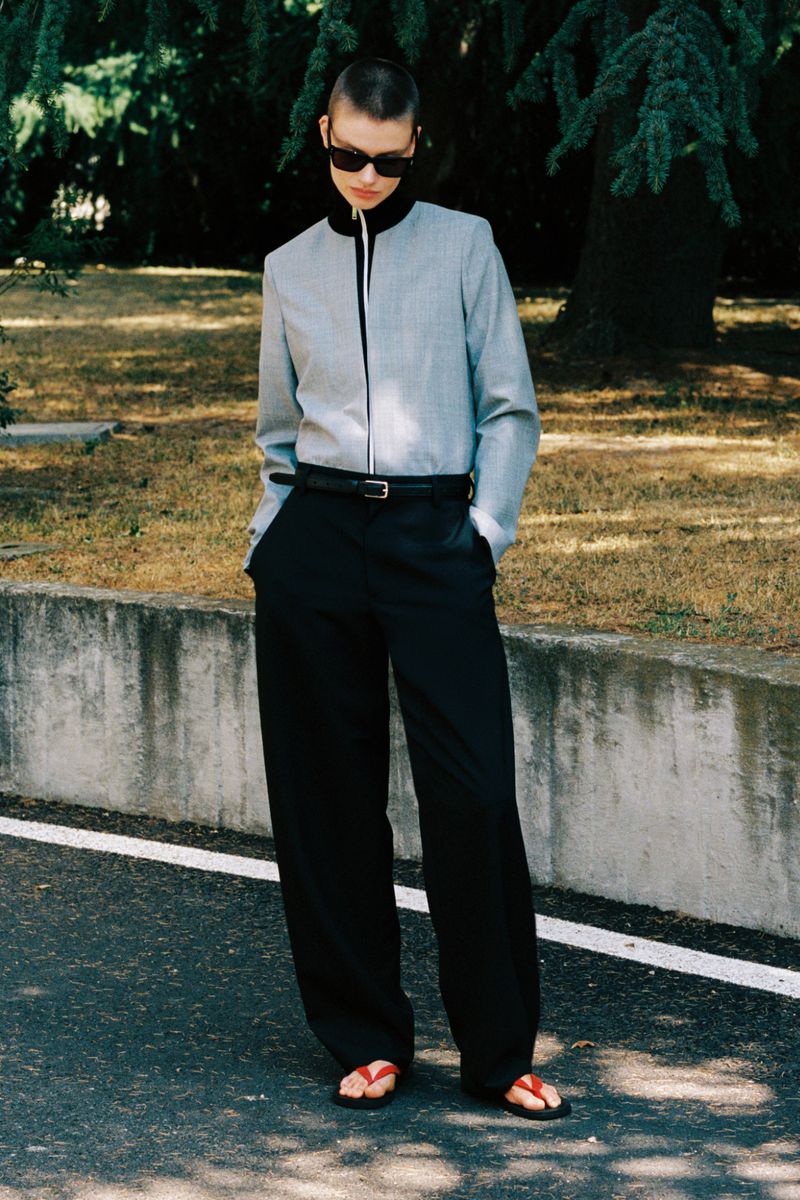 Model with short buzzcut and dark sunglasses wearing grey zip-up jacket and black trousers outdoors near a stone wall