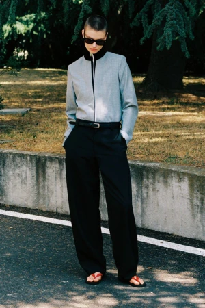 Model with short buzzcut and dark sunglasses wearing grey zip-up jacket and black trousers outdoors near a stone wall