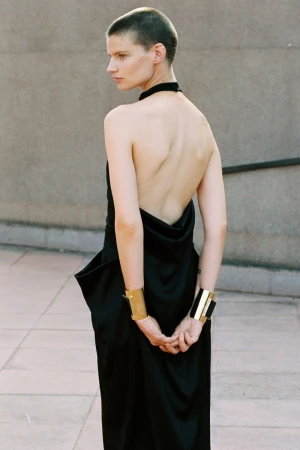 Model with shaved head wearing a backless black dress and gold cuffs outdoors