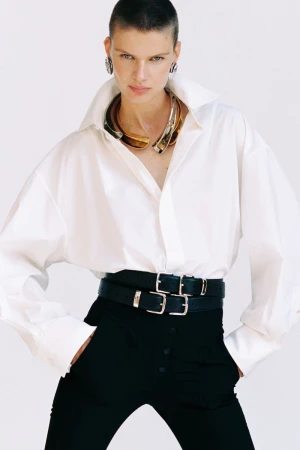 Model with short hair wearing a white collared shirt and black trousers with layered belts against a white background