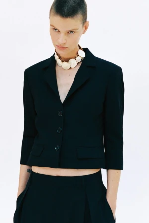 Model with short hair wearing a black buttoned blazer and a large round beaded necklace against a white background