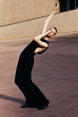 Model with shaved hair wearing a long black dress posing dramatically on a paved public square outdoors