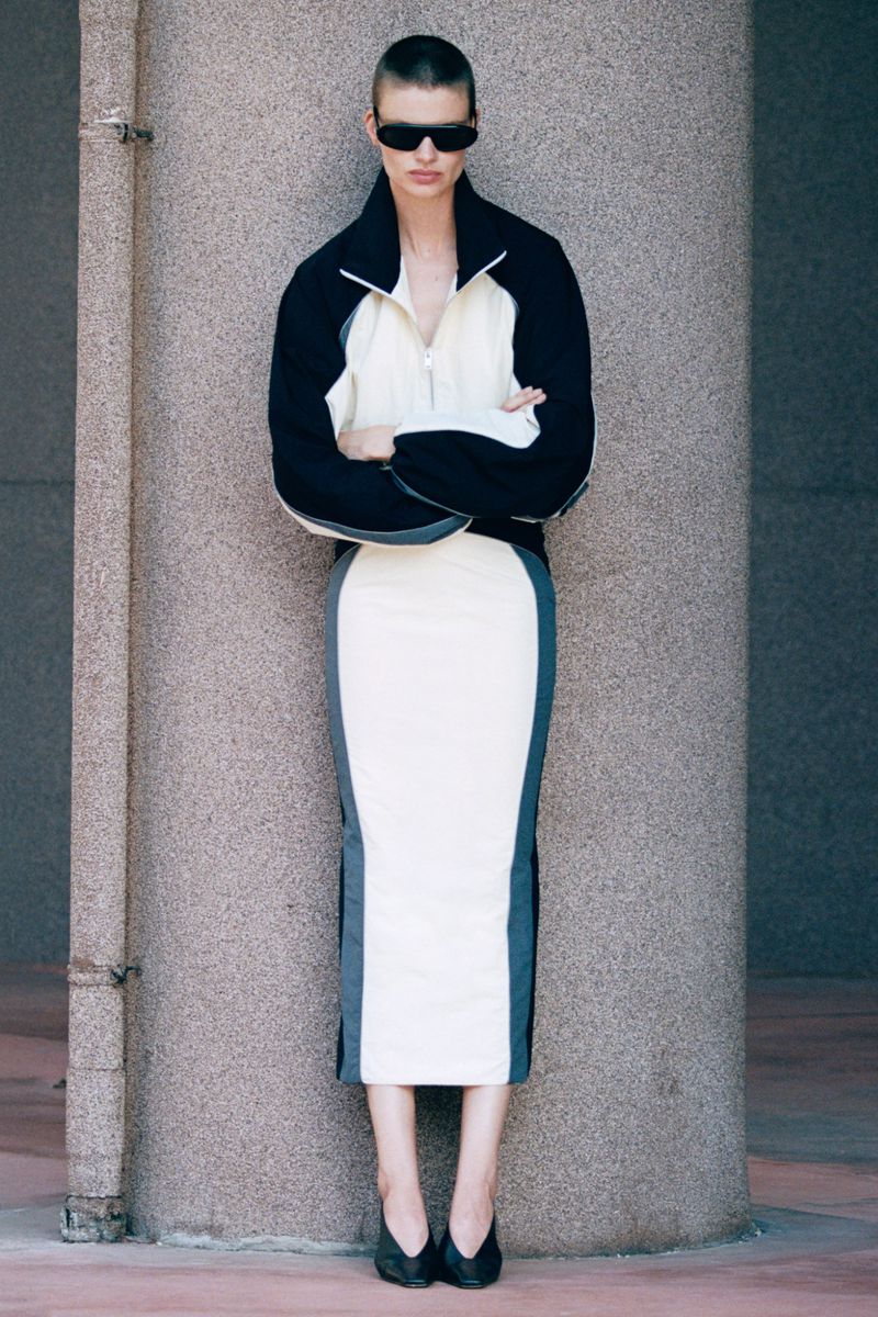 Model with short hair and sunglasses wearing a colorblocked black and white outfit leaning against a concrete pillar