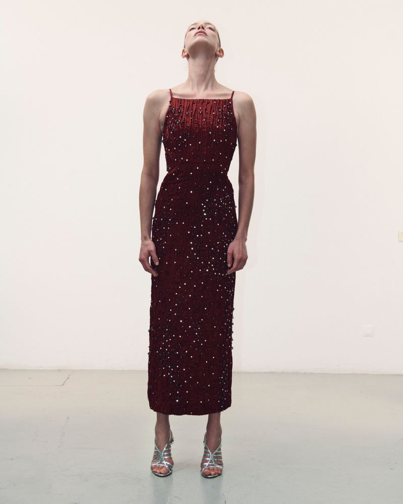 Model standing straight with head tilted back wearing a dark red embellished dress and silver heels on plain white backdrop