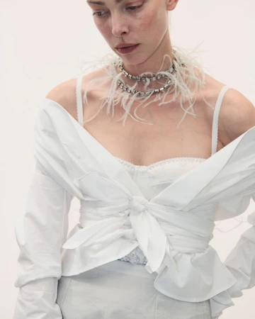 Model wearing a white tied shirt with a feather and rhinestone necklace against a white background